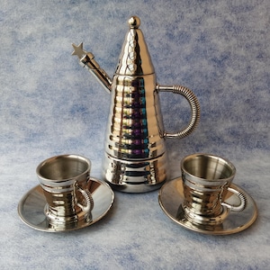 Puede incluir: Una cafetera espresso plateada con un adorno en forma de estrella en el pico, acompañada de dos tazas y platillos a juego. El juego tiene una textura martillada y un acabado brillante.