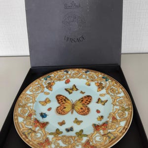 May include: A decorative plate with a light blue background and a gold rim. The plate features a large orange and yellow butterfly in the centre, surrounded by smaller butterflies and ladybirds. The plate is in a black box with the Versace logo.