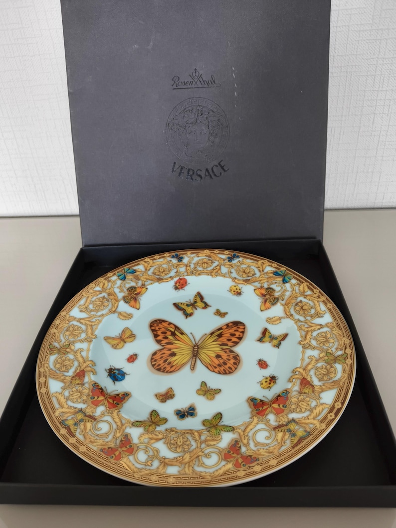 May include: A decorative plate with a light blue background and a gold rim. The plate features a large orange and yellow butterfly in the centre, surrounded by smaller butterflies and ladybirds. The plate is in a black box with the Versace logo.