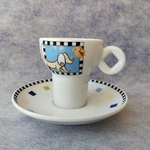 May include: White espresso cup and saucer set. The cup features a cartoon dog design on a blue background, with a square handle. The saucer has a black and white checkered border with colorful square accents.