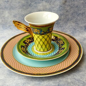 May include: A decorative teacup and saucer set. The teacup has a gold handle and a yellow, green, and blue patterned design. The set includes a pink plate with a gold border, a green plate, and a teal plate.