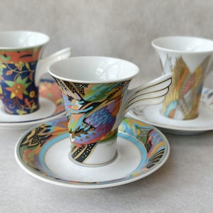 May include: Three decorative ceramic teacups and saucers. Each cup has a unique colorful design, with wing-shaped handles. The saucers and cups feature floral and abstract patterns in shades of blue, orange, and gold. The cups are on a white background.