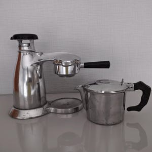 Puede incluir: Una cafetera espresso manual vintage hecha de metal plateado pulido con mangos de plástico negro y una jarra de metal plateado a juego. La cafetera cuenta con una caldera vertical, un brazo portafiltro y una base circular de apoyo.