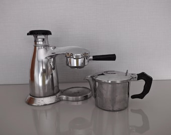 Op de afbeelding: Een vintage handmatige espressomachine van gepolijst zilverkleurig metaal met zwarte kunststof handgrepen en een bijpassende zilverkleurige metalen kan. De espressomaker heeft een verticale boiler, een filterdragerarm en een ronde voet.