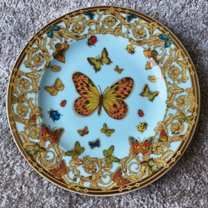 May include: A decorative plate with a light blue centre and a brown rim. The plate features a gold baroque design and colourful butterflies. The butterflies are in various sizes and colours, including orange, yellow, blue, and red.