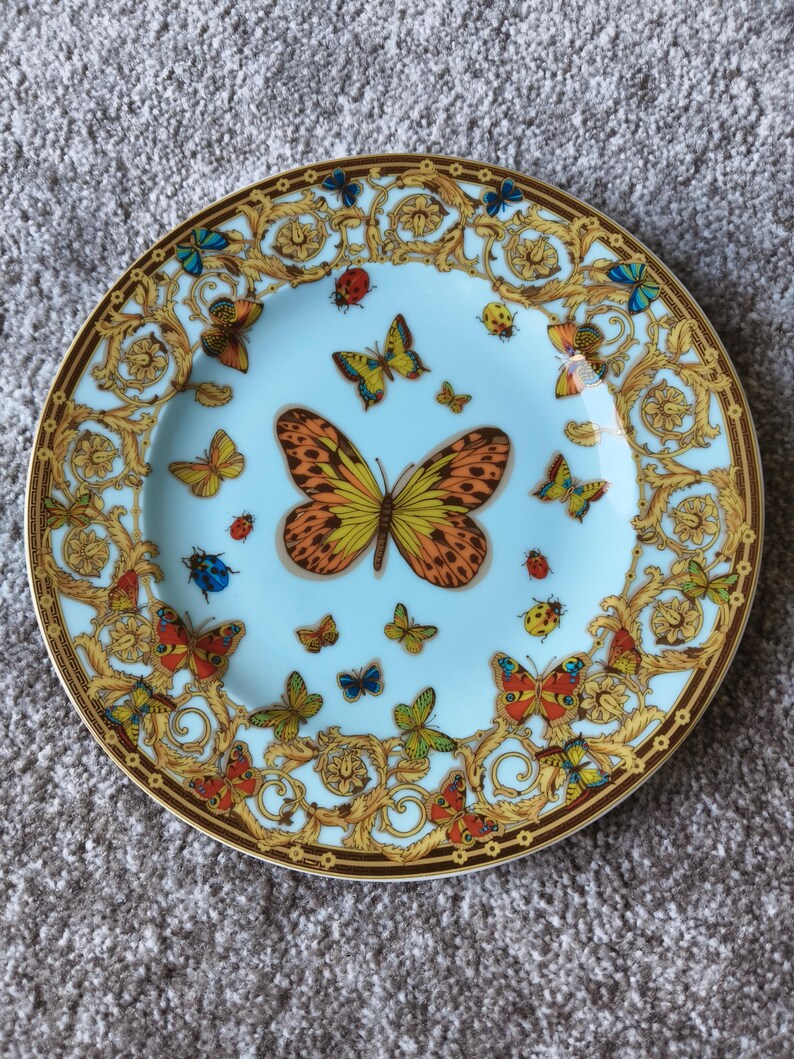 May include: A decorative plate with a light blue centre and a brown rim. The plate features a gold baroque design and colourful butterflies. The butterflies are in various sizes and colours, including orange, yellow, blue, and red.