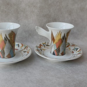 May include: Two white ceramic espresso cups with matching saucers. The cups have a unique wing-shaped handle and are decorated with colorful abstract designs in shades of orange, yellow, and blue. The saucers feature a complementary pattern.
