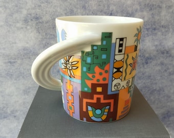 May include: White ceramic mug with a large, curved handle. The mug is decorated with a colourful geometric pattern featuring shapes, suns, and faces in shades of orange, green, blue, and brown. The mug sits on a dark grey surface.