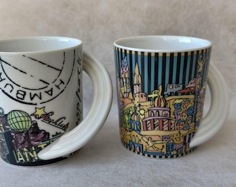 May include: Two white ceramic mugs with distinctive curved handles. One mug displays the word "HAMBURG" and a cityscape. The other mug has a colourful cityscape design with vertical blue stripes.