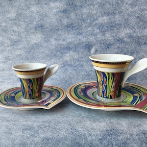 May include: Two decorative espresso cups and saucers. The cups have a white interior and a colorful exterior with yellow, blue, red, and green patterns. The saucers have a matching design. The handles are white and curved.