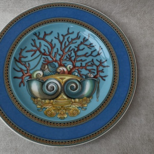 May include: A decorative plate with a blue border and a central design featuring coral, seashells, and a stylized shell-shaped element. The plate has a light blue center and gold accents.