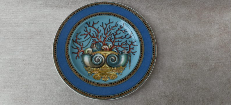 May include: A decorative plate with a blue border and a central design featuring coral, seashells, and a stylized shell-shaped element. The plate has a light blue center and gold accents.