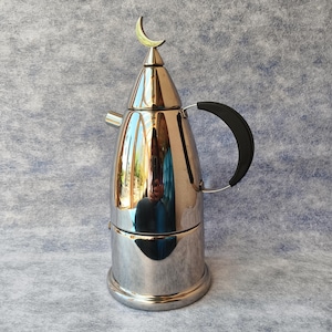 May include: A shiny, stainless steel coffee pot with a black handle and a crescent moon-shaped accent on top. The pot has a rounded body and a spout. The pot is reflective, showing the surrounding environment.