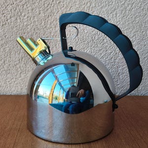 May include: A stainless steel kettle with a black handle and a gold-colored spout. The kettle has a reflective surface, and the handle is arched. The spout is a unique design. The kettle is sitting on a wooden surface.
