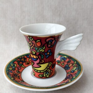 May include: A decorative espresso cup and saucer set. The cup and saucer feature a vibrant floral design in red, yellow, blue, and green. The cup has a unique wing-shaped handle. The saucer has a green border.