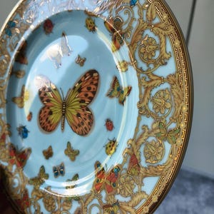 May include: A decorative plate with a pale blue background and a gold rim. The plate features colourful butterflies and gold floral patterns. The plate has a glossy finish, likely made of porcelain.