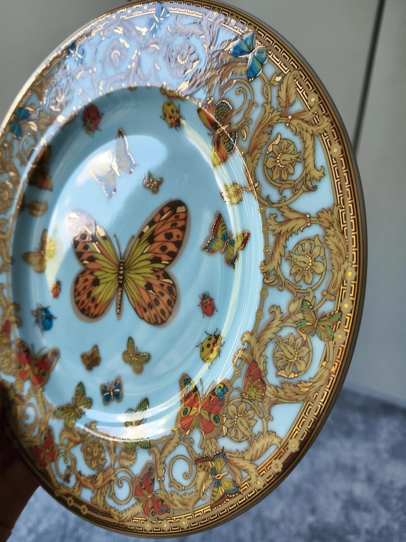 May include: A decorative plate with a pale blue background and a gold rim. The plate features colourful butterflies and gold floral patterns. The plate has a glossy finish, likely made of porcelain.