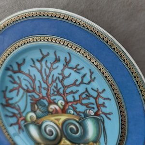May include: Decorative plate with a blue rim and a central design featuring coral and seashells. The plate has a gold trim and a light blue center. The coral is reddish-brown, and the seashells are teal.