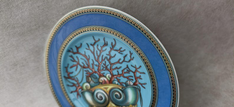 May include: Decorative plate with a blue rim and a central design featuring coral and seashells. The plate has a gold trim and a light blue center. The coral is reddish-brown, and the seashells are teal.