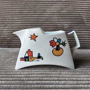 May include: White ceramic creamer with colorful abstract illustrations. The creamer features a star, house, and abstract flower design. The handle is white with a blue accent. The creamer has a unique, angular shape.