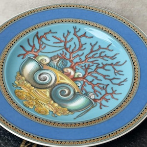 Puede incluir: Plato decorativo con borde azul y diseño central con conchas marinas, coral y detalles dorados. El plato tiene un centro turquesa y un borde dorado y blanco. El plato es probablemente de cerámica o porcelana.