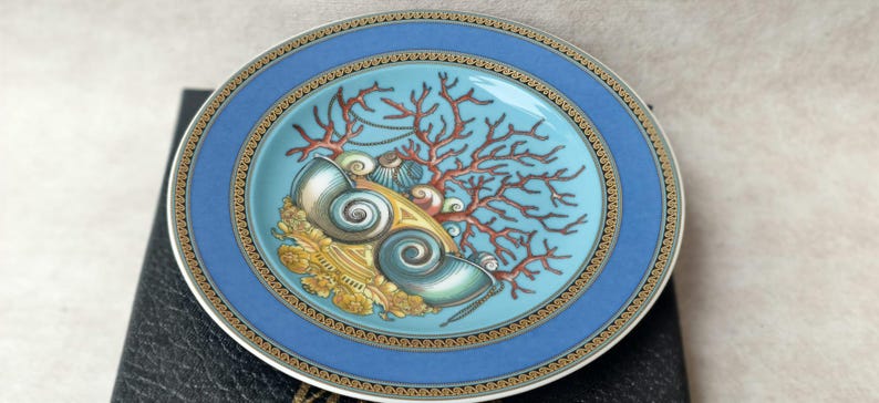 May include: Decorative plate with a blue border and a central design featuring seashells, coral, and gold accents. The plate has a turquoise center and a gold and white rim. The plate is likely ceramic or porcelain.