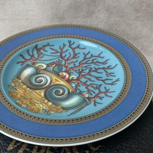 May include: A decorative plate with a blue border and a central design featuring seashells, coral, and gold accents. The plate has a turquoise center and a gold and brown patterned border. The plate is round and appears to be made of ceramic.
