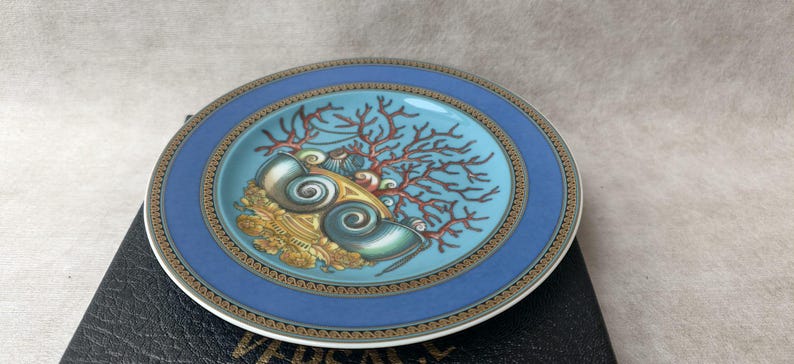 May include: A decorative plate with a blue border and a central design featuring seashells, coral, and gold accents. The plate has a turquoise center and a gold and brown patterned border. The plate is round and appears to be made of ceramic.