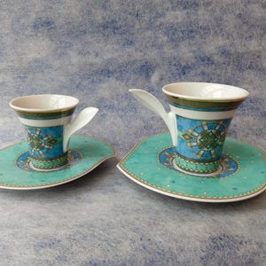May include: Two ceramic teacups and saucers. The cups have a white interior and a turquoise and blue exterior with a decorative pattern. The saucers are turquoise with a matching design. The handles are white and curved.