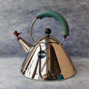 May include: A stainless steel kettle with a teal handle and a small bird-shaped whistle. The kettle has a black knob on top and a circular logo that says "ALESSI". The kettle is a polished silver color.