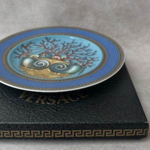 May include: A small, round decorative plate with a blue border and a central design featuring seashells and coral. The plate rests on a black box with the brand name "VERSACE" visible. The plate's design includes gold and teal accents.