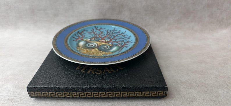 May include: A small, round decorative plate with a blue border and a central design featuring seashells and coral. The plate rests on a black box with the brand name "VERSACE" visible. The plate's design includes gold and teal accents.