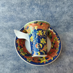 May include: A decorative teacup and saucer set. The cup and saucer feature a vibrant floral design in shades of blue, orange, yellow, and green. The cup has a unique wing-shaped handle.