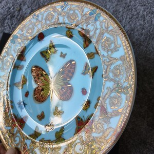 May include: A decorative plate with a light blue background and a gold rim. The plate features a large butterfly in the centre, surrounded by smaller butterflies and ladybirds. Gold floral patterns adorn the rim.