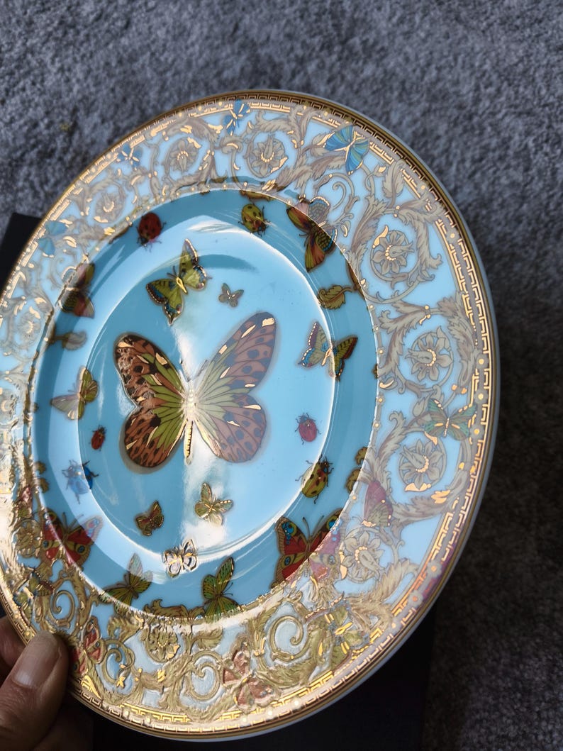 May include: A decorative plate with a light blue background and a gold rim. The plate features a large butterfly in the centre, surrounded by smaller butterflies and ladybirds. Gold floral patterns adorn the rim.