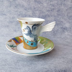May include: A white ceramic teacup and saucer set. The teacup features a blue Pegasus design and a wing-shaped handle. The saucer has a colorful geometric and floral pattern. The set is on a light blue background.
