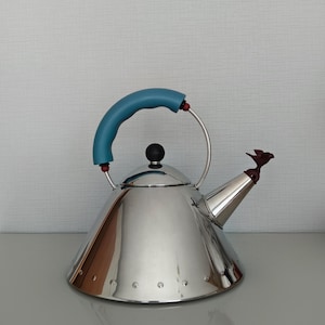 May include: A stainless steel tea kettle with a blue handle and a bird-shaped spout. The kettle has a black knob on the lid and a series of rivets around the base. The handle is arched and attached to the kettle with metal supports.