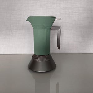 May include: A modern, two-tone espresso maker. The upper portion is a muted green with vertical ridges, while the base and handle are a metallic gray. The spout is on the left side. The coffee maker sits on a reflective surface.