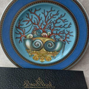 May include: A decorative plate with a blue border and a central design featuring coral, seashells, and a decorative bowl. The plate is set on a surface next to a black leather case with gold lettering.