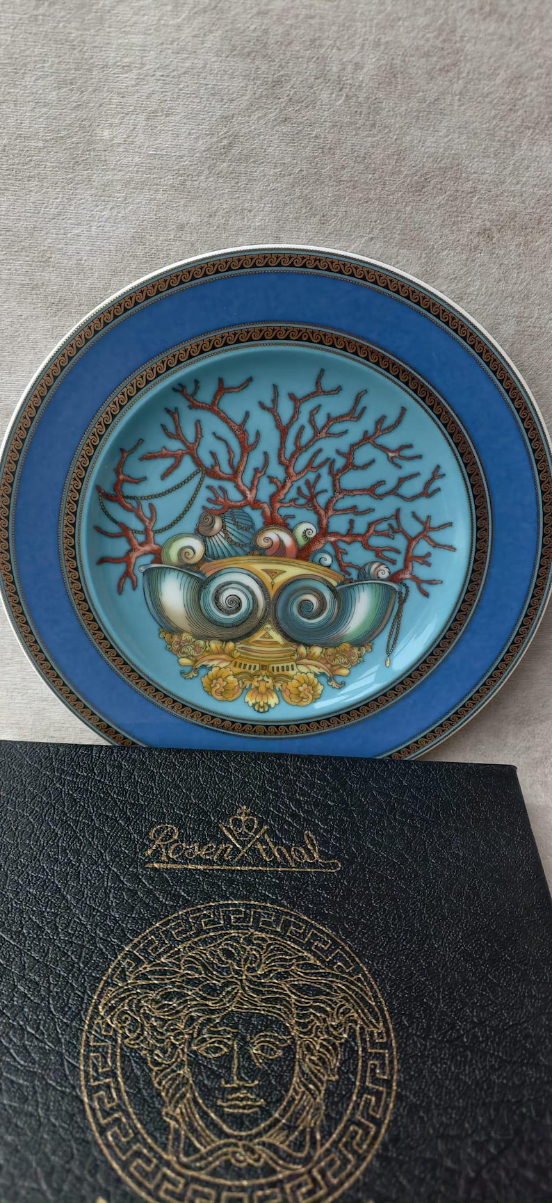 May include: A decorative plate with a blue border and a central design featuring coral, seashells, and a decorative bowl. The plate is set on a surface next to a black leather case with gold lettering.