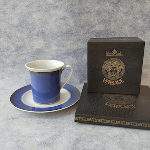 May include: A blue and white porcelain teacup and saucer set with a Greek key design. The teacup and saucer are on a light blue background. A black box with the Versace logo is also in the image.