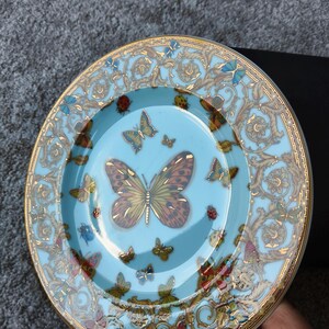 May include: A decorative plate with a light blue centre and a gold rim. The plate features a butterfly design with various butterflies in different sizes and colours. The rim has a gold ornate pattern.