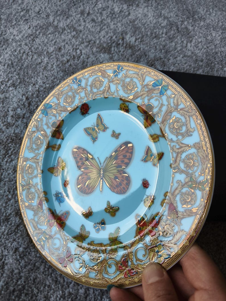 May include: A decorative plate with a light blue centre and a gold rim. The plate features a butterfly design with various butterflies in different sizes and colours. The rim has a gold ornate pattern.