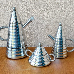 Op de afbeelding: Een set van drie gepolijste zilveren thee- of koffieserviesstukken. De set bevat een hoge, conische theepot met een stervormig tuitdetail, een kleinere theepot en een suikerpot, allemaal met een getextureerd oppervlak en afgeronde handvatten.