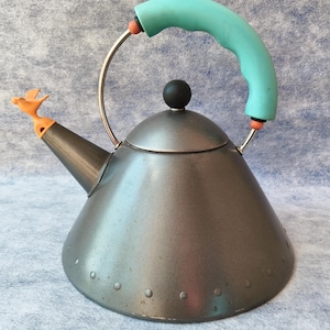 May include: A gray metal kettle with a teal handle and a small orange bird on the spout. The kettle has a black knob on the lid and a silver handle. The base of the kettle has a series of small rivets.