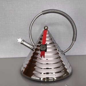 May include: A polished stainless steel conical tea kettle with a ribbed texture, a coiled metal handle, and a spout featuring a star-shaped tip. A red ribbon is tied to the top knob, and a black tag with a star logo is attached to the side.