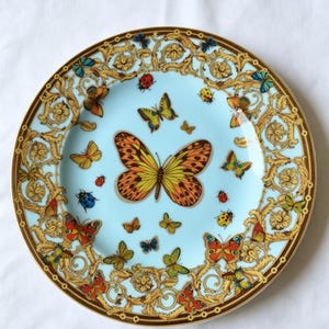 May include: A decorative plate with a light blue centre, adorned with butterflies in orange, yellow, red, and blue. The plate has a gold and brown ornate border with additional butterfly and ladybug designs.