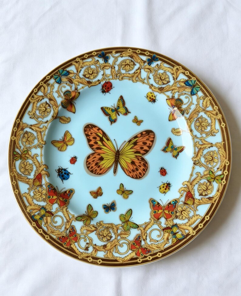 May include: A decorative plate with a light blue centre, adorned with butterflies in orange, yellow, red, and blue. The plate has a gold and brown ornate border with additional butterfly and ladybug designs.