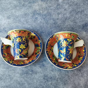 May include: Two matching teacups and saucers. The cups and saucers have a vibrant design with a blue background, orange rim, and floral patterns in yellow, green, and white. The handles are white. The set is placed on a blue surface.