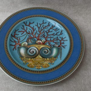 May include: A decorative plate with a blue border and a central design featuring coral, seashells, and a stylized shell-like structure. The plate has a gold and black patterned trim and a light blue center.